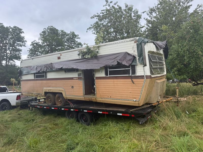 Derelict travel trailer with a torn tarp loaded on a flatbed behind the AllStarr Solutions truck
