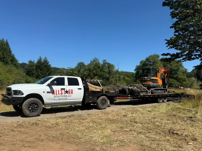 AllStarr Solutions truck towing an excavator on a flatbed trailer down a gravel track